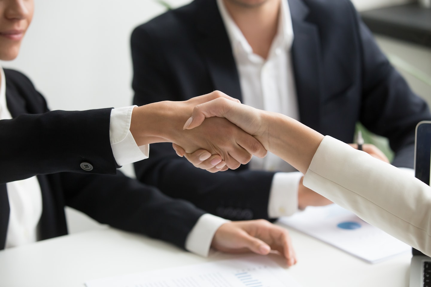 Two business professionals shake hands over a desk in an office.